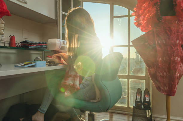 woman sitting in her atelier using the sewing machine by the sunlit window caucasian woman sitting in her atelier concentrated using the sewing machine by the sunlit window, designing a fashion red color dress. concentrated solar power photos stock pictures, royalty-free photos & images