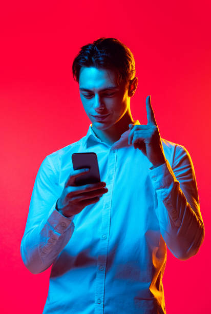 portrait of young man, student looking on phone and rising finger up isolated over red studio background in blue neon light - blue vertical color image photography imagens e fotografias de stock
