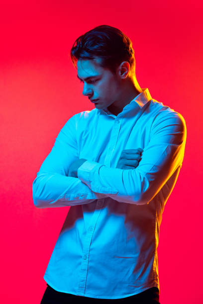 портрет молодого красавца в белой рубашке, смотрящего вниз, позирующего изолированно на красном студийном фоне в синем неоновом цвете - red blue white neon light стоковые фото и изображения