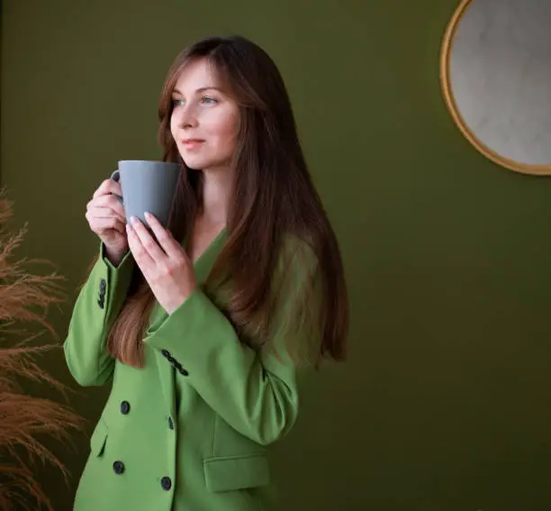 Pretty adult girl with long brown hair wearing green suit,jacket, drinking tea or coffee .Business young woman holding grey mug,cup.