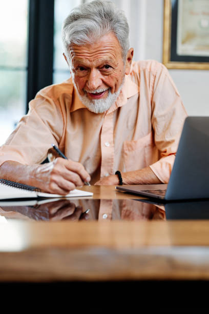 istock A happy senior student is sitting at home and writing down notes in a notebook while following an online lecture on the laptop. 1380578311