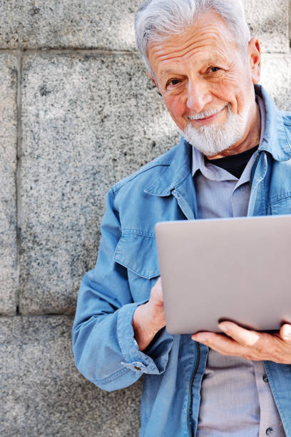 istock A happy senior student is standing against the wall and typing on a laptop. 1380578716