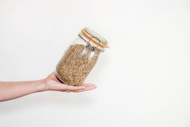 A hand holding a glass jar full of lentils. Concept of storage of legumes in the pantry. Vegetal protein. White background.