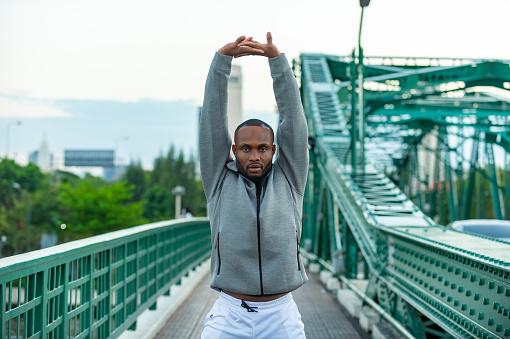 Confidence African man in sportswear stretching his body while jogging exercise on the bridge in the city at summer sunset. Strong male athlete enjoy outdoor activity sport training running workout