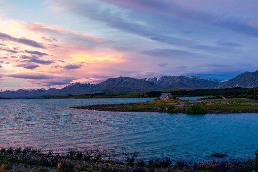 This November 2021 sunset image shows a wide-shot of the historic Church of the Good Shepherd in Lake Tekapo, Aotearoa New Zealand. The sunset illuminates storm clouds, providing a dramatic sky. Wind whips up the lake. Blooming lupins are seen along the lakeshore. The snowcapped Te Tiritiri-o-te-moana Southern Alps are in the left background, while Mount Gerald is in the centre background.