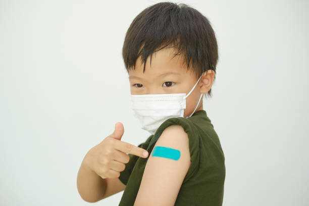 Little doubtful kid wearing medical mask showing his arm with bandage after receiving vaccination stock photo