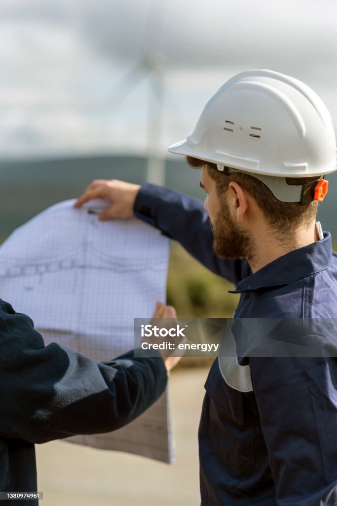 Young man engineers holding blueprints and checking wind turbines on site. Discussion Stock Photo