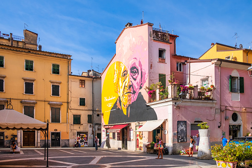 In the Piazzetta delle Erbe of Carrara stands out the mural titled 'Non abbandonare la città' (Do not abandon the city), a work of the artistic duo Orticanoodles dated 2013 and dedicated to Francesca Rolla, one of the heroines of the Second World War. People in the square.