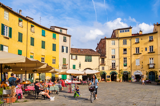 People sitting at the outdoor tables the Piazza dell'Anfiteatro of Lucca, an elliptical shape square surrounded by a ring of buildings that follows the former second century Roman amphitheater of the city.