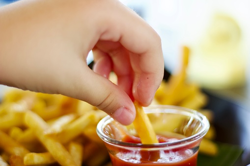 Male child hand dipping french fries in a tomato ketchup sauce close up shot, under junk food with children health concept of use