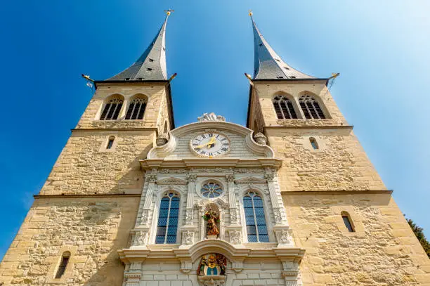 Frog's perspective of the front of the Catholic Court Church of St. Leodegar in Lucerne with its two towers and a clock with dial in sunshine and cloudless sky