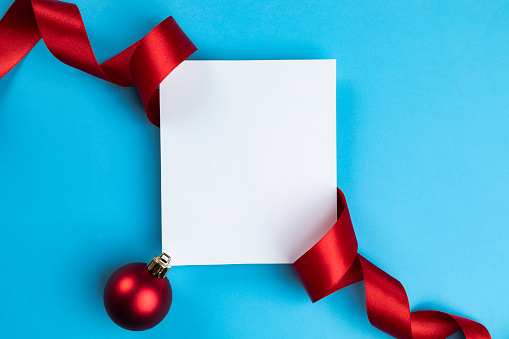 White blank card on blue background wrapped with red ribbon. At the left bottom corner of the blank card is one red christmas bauble.