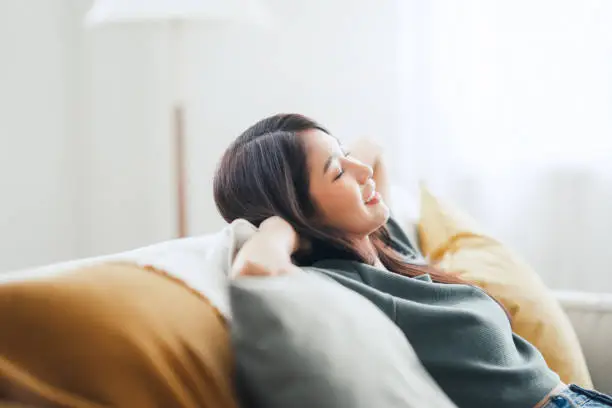 Photo of Relaxed young asian woman enjoying rest on comfortable sofa at home, calm attractive girl relaxing and breathing fresh air in home