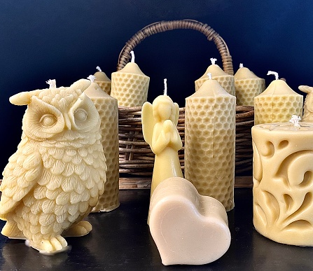 Horizontal close up of handmade beeswax candles of owl heart and other designs against black background with wicket basket