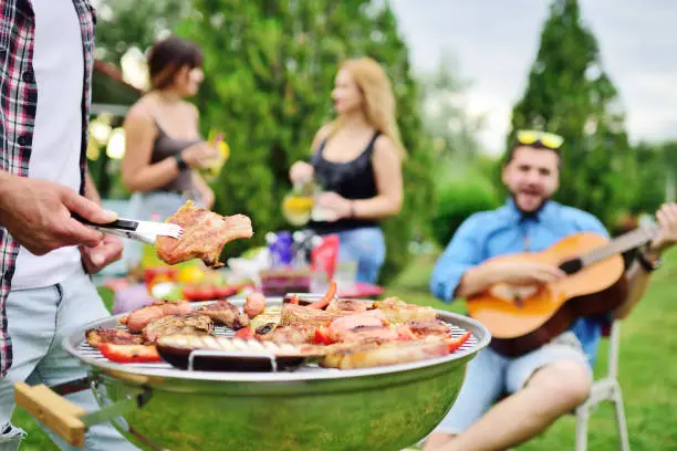 Photo of man's hand holds a barbecue tongs with a juicy delicious meat steak against the background of a barbecue grill with meat and vegetables and a group of friends on a picnic who are having fun and playing the guitar