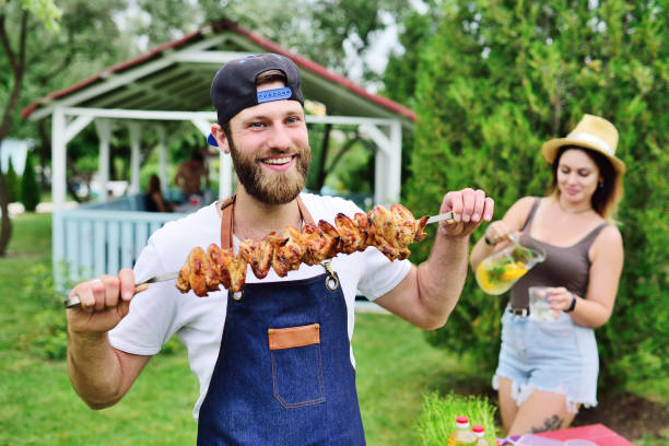 young bearded man in a cap and denim apron smiles and holds a skewer with delicious barbecue meat cooked on the grill against the background of greenery, a Park and a gazebo stock photo