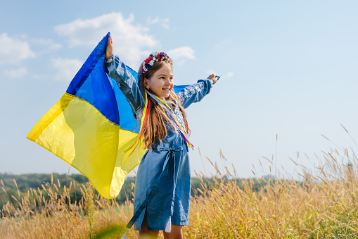 Portrait of little Ukrainian girl wearing national dress and wreath of flowers holding flag of Ukraine looking forward. A symbol of faith in peace for all people of Ukraine.