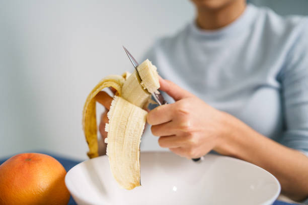 istock Caucasian young woman peeling fruit while sitting by the table at home - front view on pretty girl holding knife and banana preparing meal - healthy eating concept copy space 1381299746