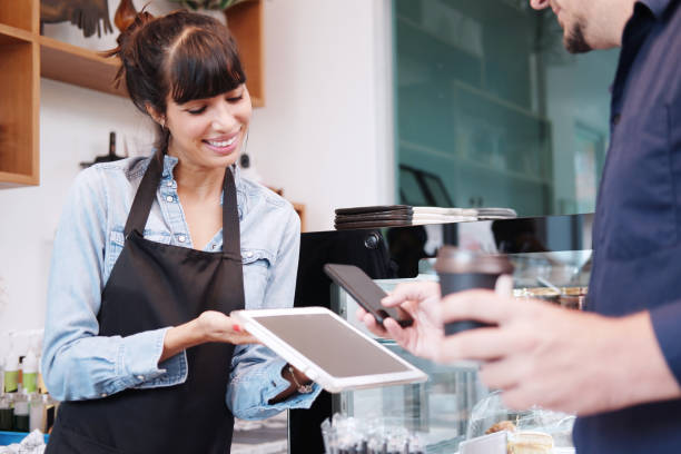 istock Young Caucasian barista woman is holding tablet for customer using smart phone scan QR code on tablet for payment at counter bar at coffee shop. Technology of digital pay without money concept. 1381311548