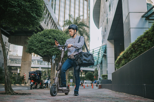 Asian Chinese male delivery person checking order address on the move with electric push scooter as mode of transport in city