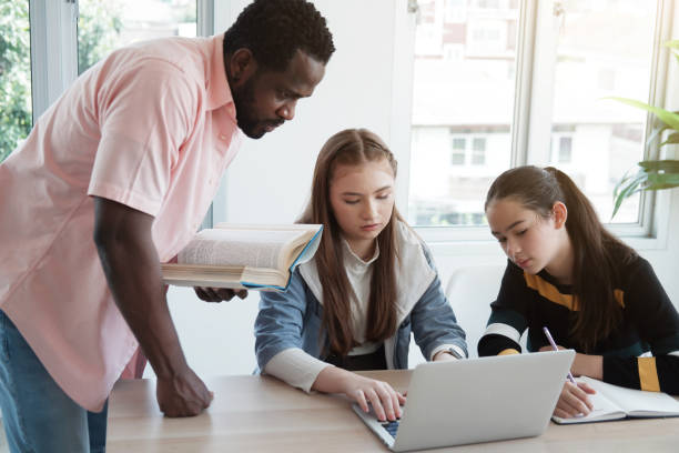 istock Young African teacher or tutor is holding book and teaching for knowledge about online research with internet on laptop for Caucasian student girls in classroom. Education and Technology Concept. 1381317447