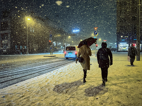 Bergen, Norway, February 19, 2022: Snow storm over the city during a winter night, while city life proceeds as normal on February 19, 2022 in Bergen, Norway. Two woman walk in the city street resisting to the heavy snow storm