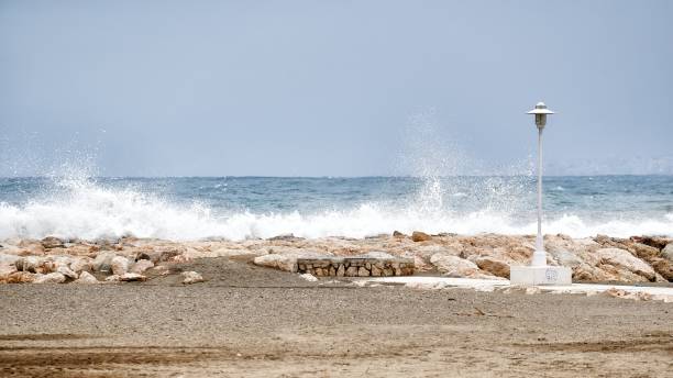 onde del mare che si infrangono duramente contro un frangiflutti vicino alla spiaggia con un lampione nelle vicinanze - sea defence concrete foto e immagini stock