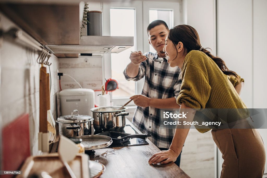 Man cooking for wife Two people, modern Japanese couple together in kitchen at home. Husband is cooking for his wife, wife is tasting it. Cooking Stock Photo