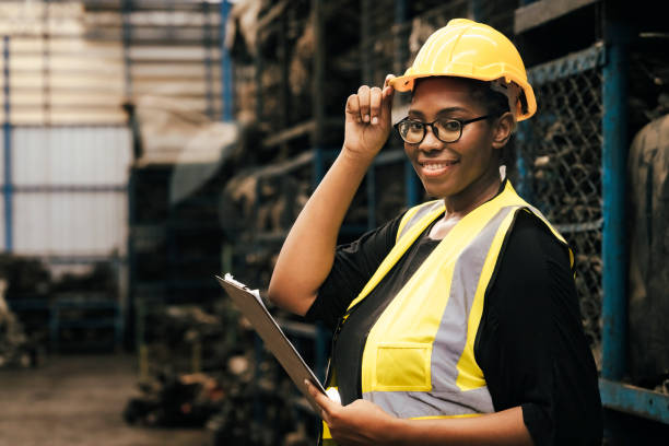 Female worker holding a hat and looking at the camera. Engineers or technicians are inspecting auto parts in warehouses and factories. African American woman holding a flip chart in parts warehouse. stock photo