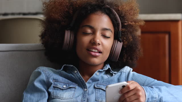 African teenage girl wear wireless headphones holds cellphone listens music