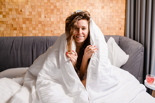 Attractive positive young woman smiling while sitting on the bed and covering her head with a blanket.