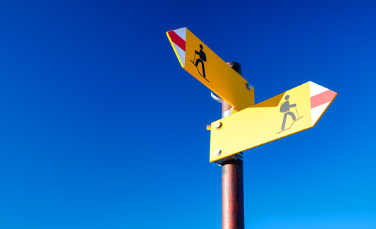 Directional marker for a mountain hike. Yellow bright sign with an arrow against the blue sky. Navigation in a hiking trip. High resolution photo.