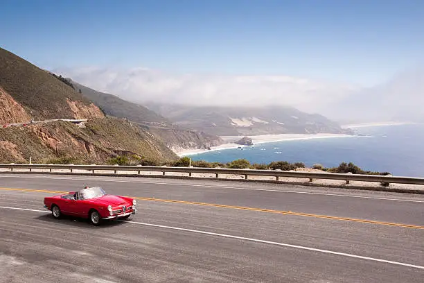A classic red convertible drives down the Pacific coast in California under a blue sky next to the ocean.