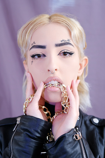 Cyberpunk blonde woman model in a black leather jacket playing with a gold chain in a studio against a pink background