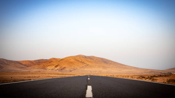 strada deserta con montagne sullo sfondo in un paesaggio arido sulle isole canarie di fuerteventura - arid climate asphalt barren blue foto e immagini stock