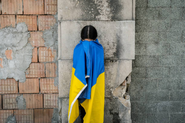 爆弾から破壊された壁の前にウクライナの旗を持つ小さな女の子。小さな女の子は平和を祈りながら国旗を振る - ukrainian culture ストックフォトと画像