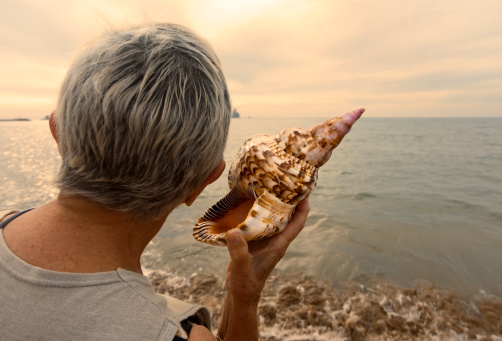 Woman listening the sound of the ocean in a conch next to the sea at dusk.