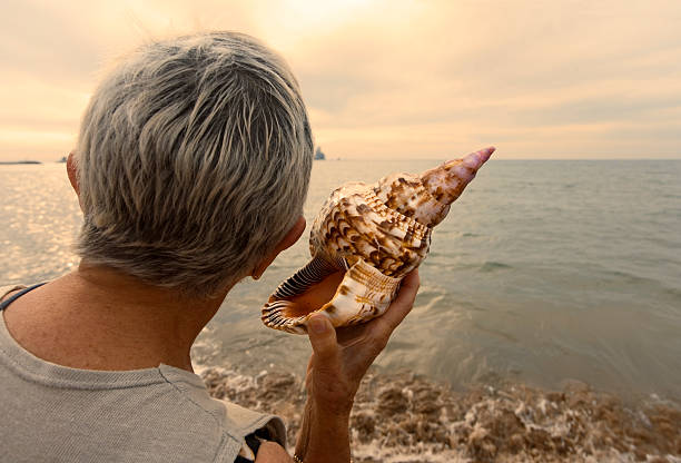 Woman holding a conch as listening the sea Woman listening the sound of the ocean in a conch next to the sea at dusk. conch shell photos stock pictures, royalty-free photos & images
