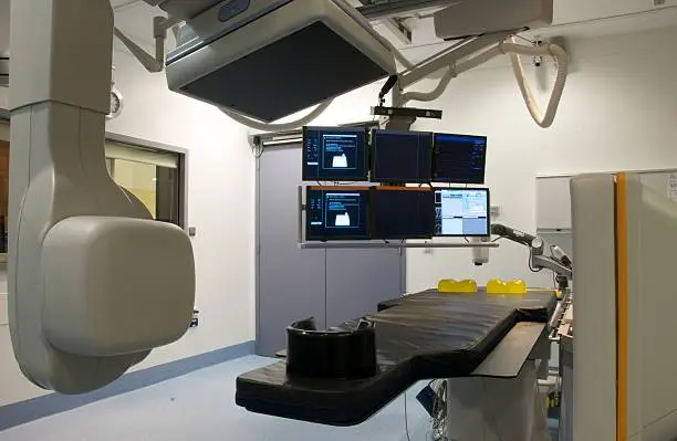 An inside view of an angiography/catheter lab with scanning equipment (x-ray and CT) in place ready for use. There are six monitors in the lab above the table the patient lies on.