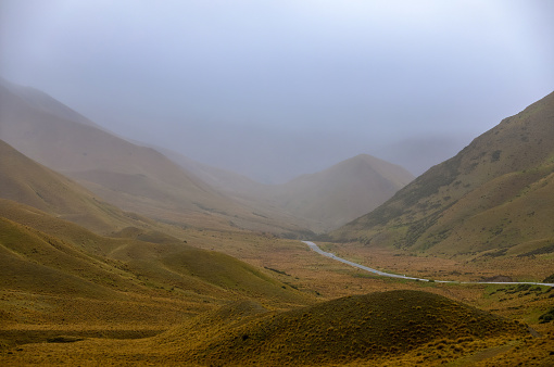 This November 2021 image shows SH 8 ascending through the Lindis Pass in Aotearoa New Zealand. The pass straddles the border of the Canterbury and Otago regions on Te Waipounamu South Island. Fog obscures part of the landscape. Tussock carpets the entire landscape.
