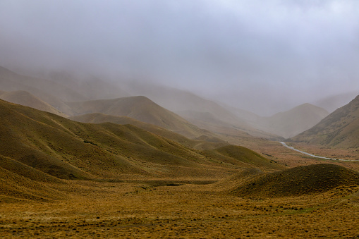 This November 2021 image shows SH 8 ascending through the Lindis Pass in Aotearoa New Zealand. The pass straddles the border of the Canterbury and Otago regions on Te Waipounamu South Island. A flowing curtain of fog obscures part of the landscape. Tussock carpets the entire landscape.