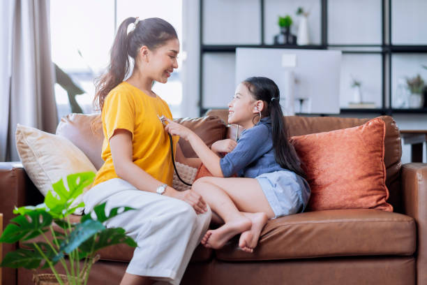 asian daughter play doctor checking up her mother with care and happiness in living room on sofa couch at home,female daughter playroll doctor nurse use stethoscope listen her mom heart pulse with fun stock photo
