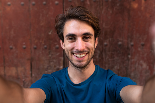 Smiling young caucasian man looking at camera taking a selfie with wood background. Lifestyle and social media concept.