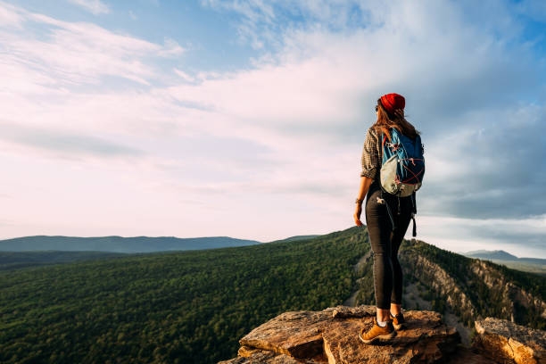 A tourist woman with a backpack admires the sunset from the top of the mountain. A traveler on the background of mountains. A tourist with a backpack is standing on the top of the mountain, panorama stock photo
