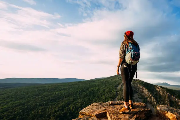 Photo of A tourist woman with a backpack admires the sunset from the top of the mountain. A traveler on the background of mountains. A tourist with a backpack is standing on the top of the mountain, panorama