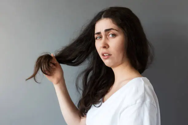 Portrait of young upset woman show dry tips of her long dark hair. Gray background. The concept of trichotillomania.