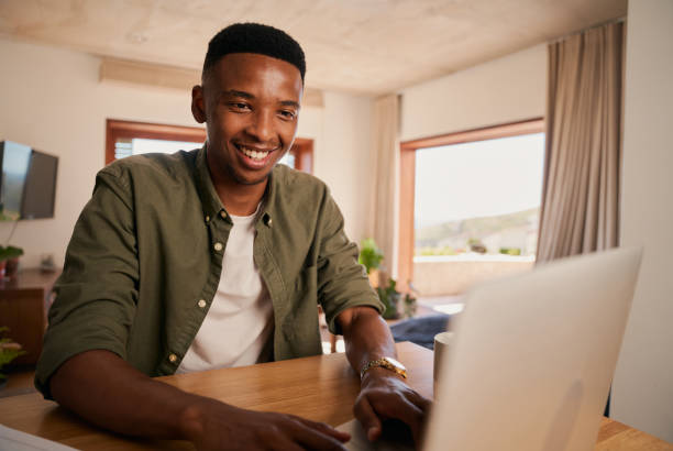 hombre negro adulto joven sonriendo mientras escribe en su computadora portátil. trabajo remoto desde su moderno apartamento - ewan fotografías e imágenes de stock