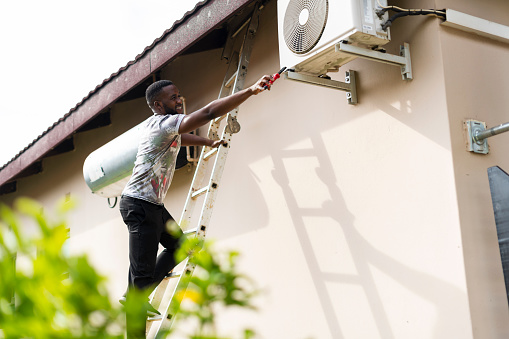Young man standing on ladder stretching dangerously with tool to fix air conditioner