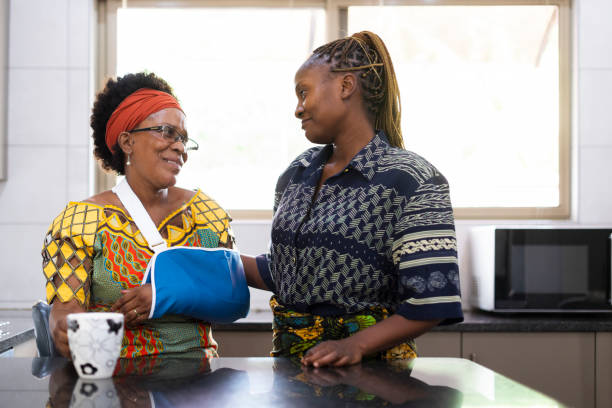 afrikanische frau mit arm in einer schlinge in küche mit erwachsener tochter - armschlinge stock-fotos und bilder