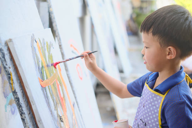 Little Asian 5 years old kindergarten boy child painting with paintbrush and watercolors on canvas stock photo